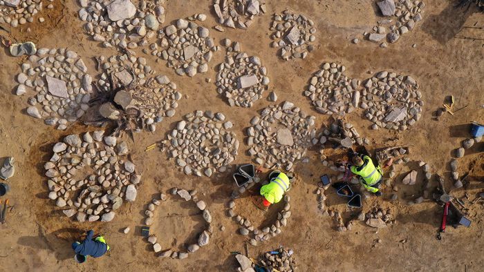Bilde av utgravingsfelt med flere sirkler laget av runde steiner. To personer i arbeidsklær jobber på feltet.